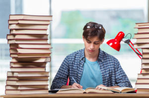 Jovem estudante de pele branca sentado estudando ao lado de duas grandes pilhas de livros